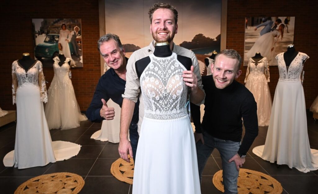 Drei Männer lächeln in einem Brautmodengeschäft, einer von ihnen trägt humorvoll ein Brautkleid. Sie sind umgeben von eleganten Hochzeitskleidern und einer fröhlichen Atmosphäre.