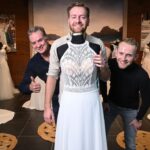 Drei Männer lächeln in einem Brautmodengeschäft, einer von ihnen trägt humorvoll ein Brautkleid. Sie sind umgeben von eleganten Hochzeitskleidern und einer fröhlichen Atmosphäre.