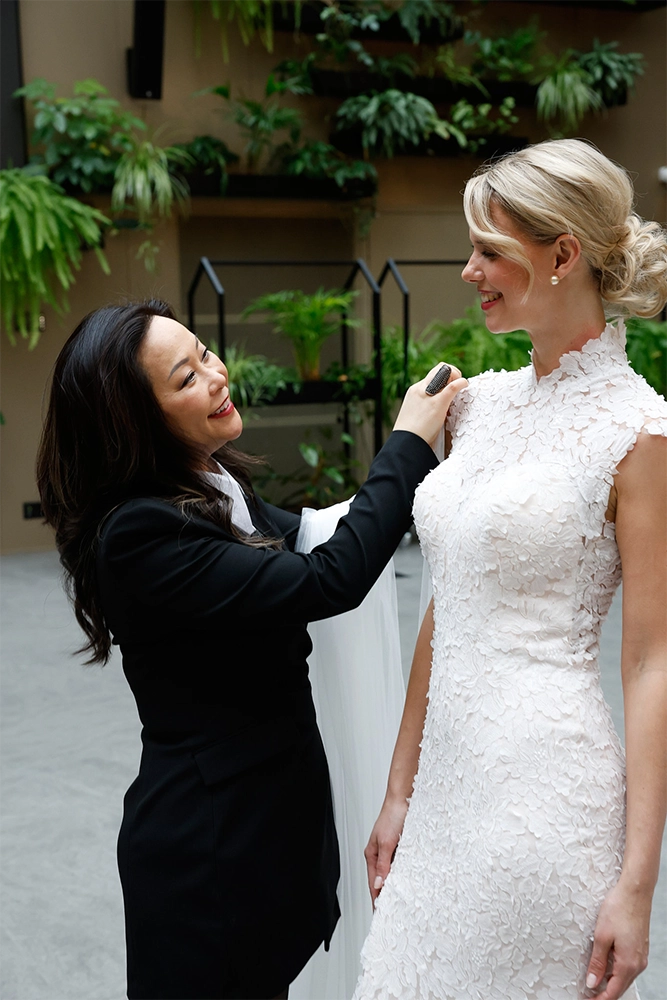Eine Designerin passt sorgfältig ein Hochzeitskleid an eine Braut in einem eleganten, begrünten Innenraum an.